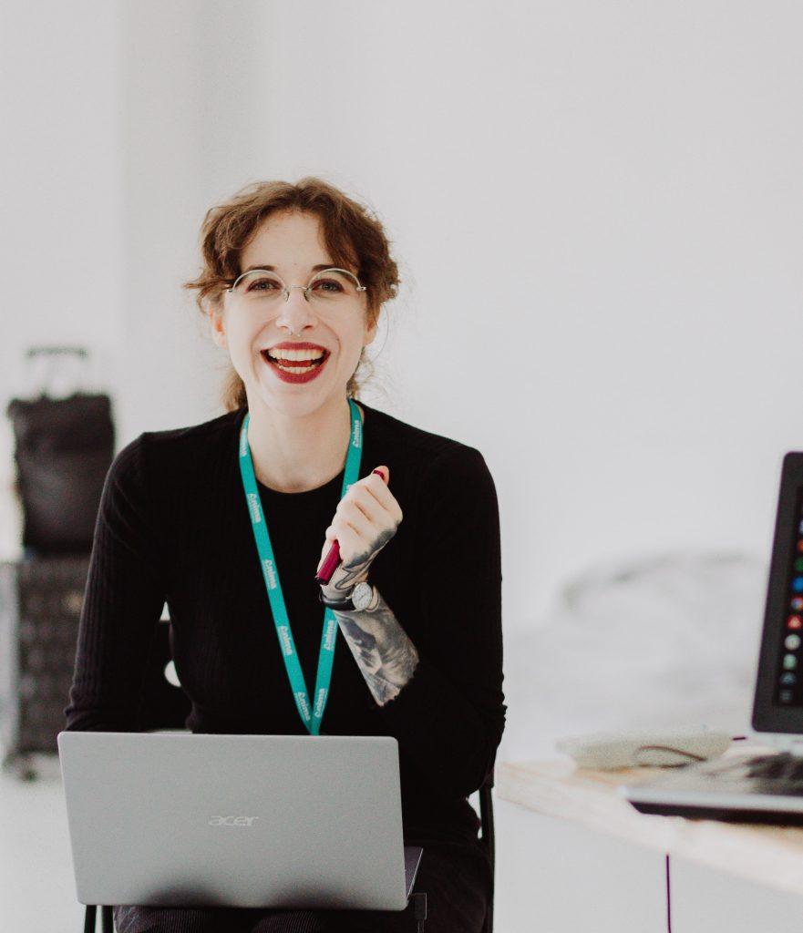 Une femme regarde la caméra en riant, assise à un bureau devant son ordinateur portable